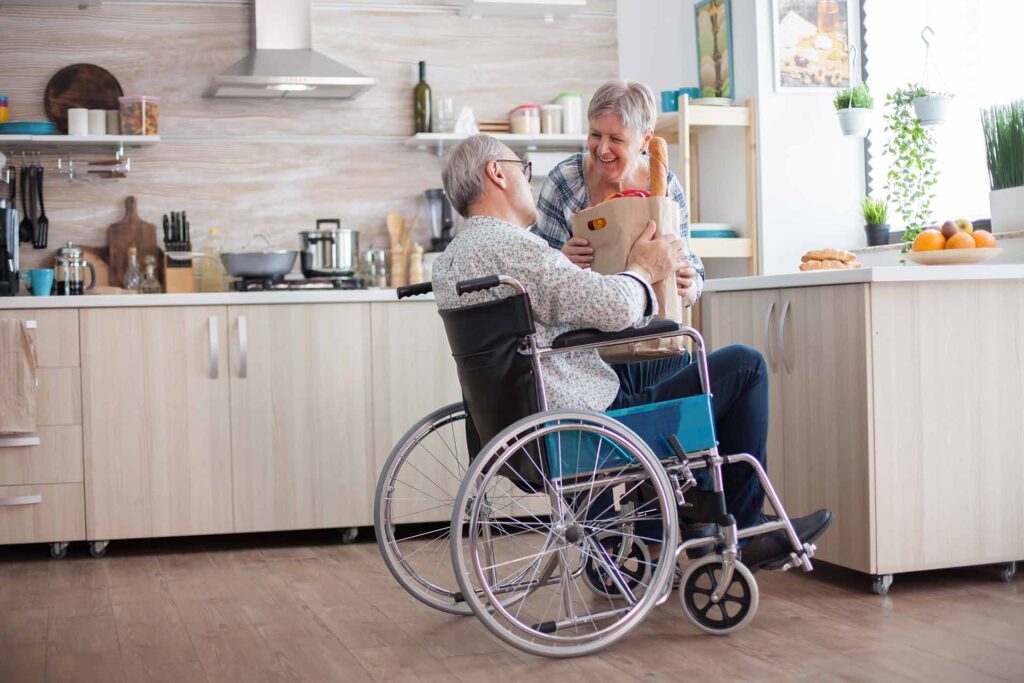 Älterer Mann im Rollstuhl reicht seiner Frau in der Küche eine Einkaufstüte